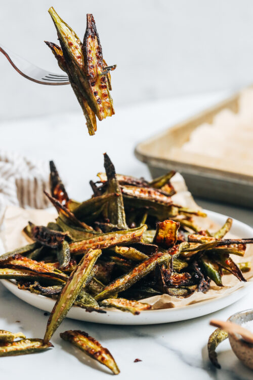 Fork holding up a few pieces of crispy oven roasted okra over a plate with more okra