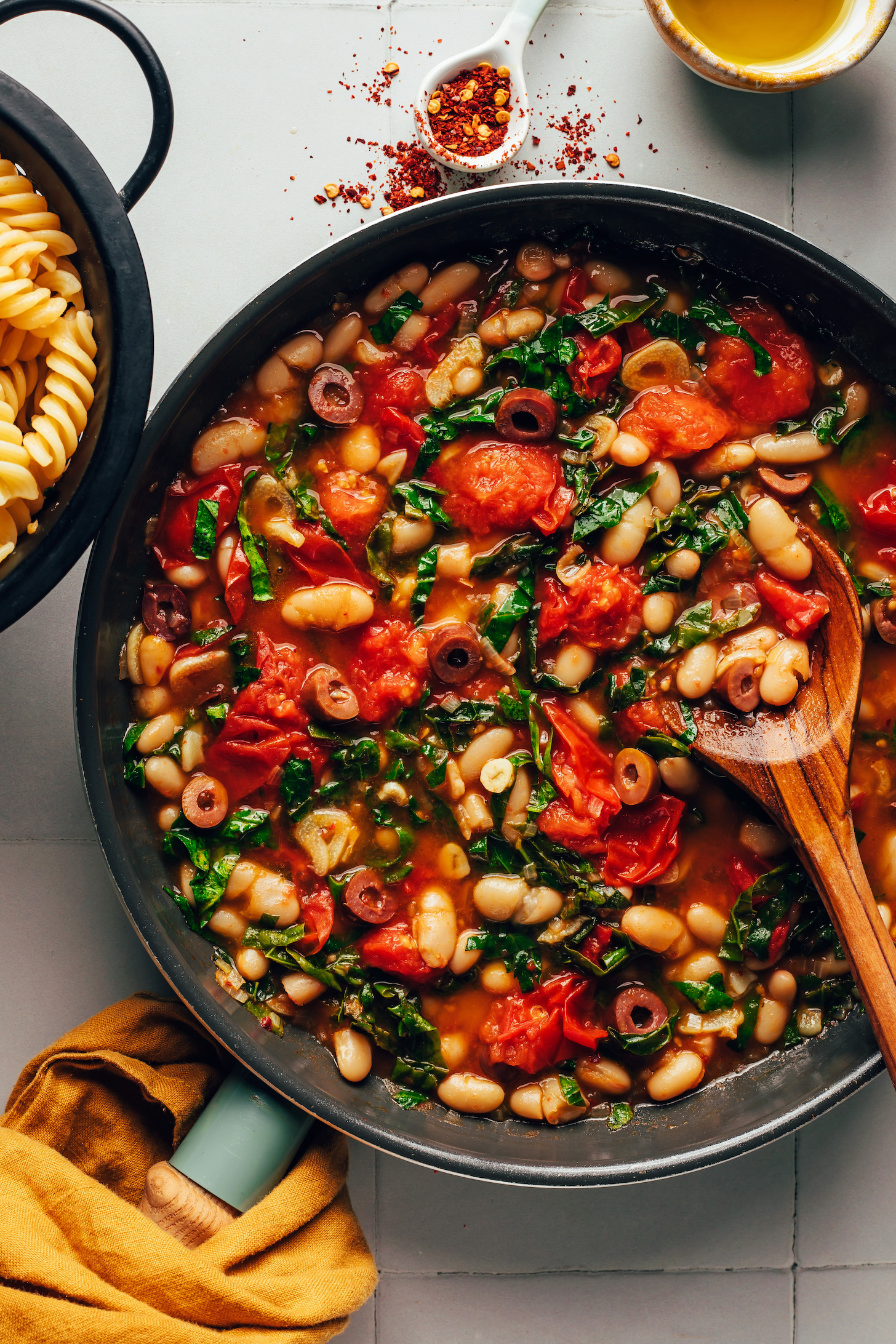 Skillet with a saucy mixture of burst cherry tomatoes, white beans, kale, olives, and seasonings
