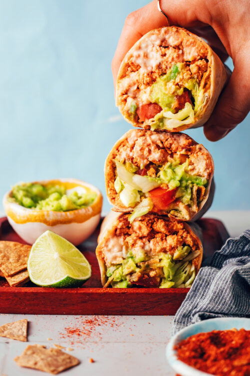 Holding a vegan crunchwrap-inspired burrito above a stack of more burritos