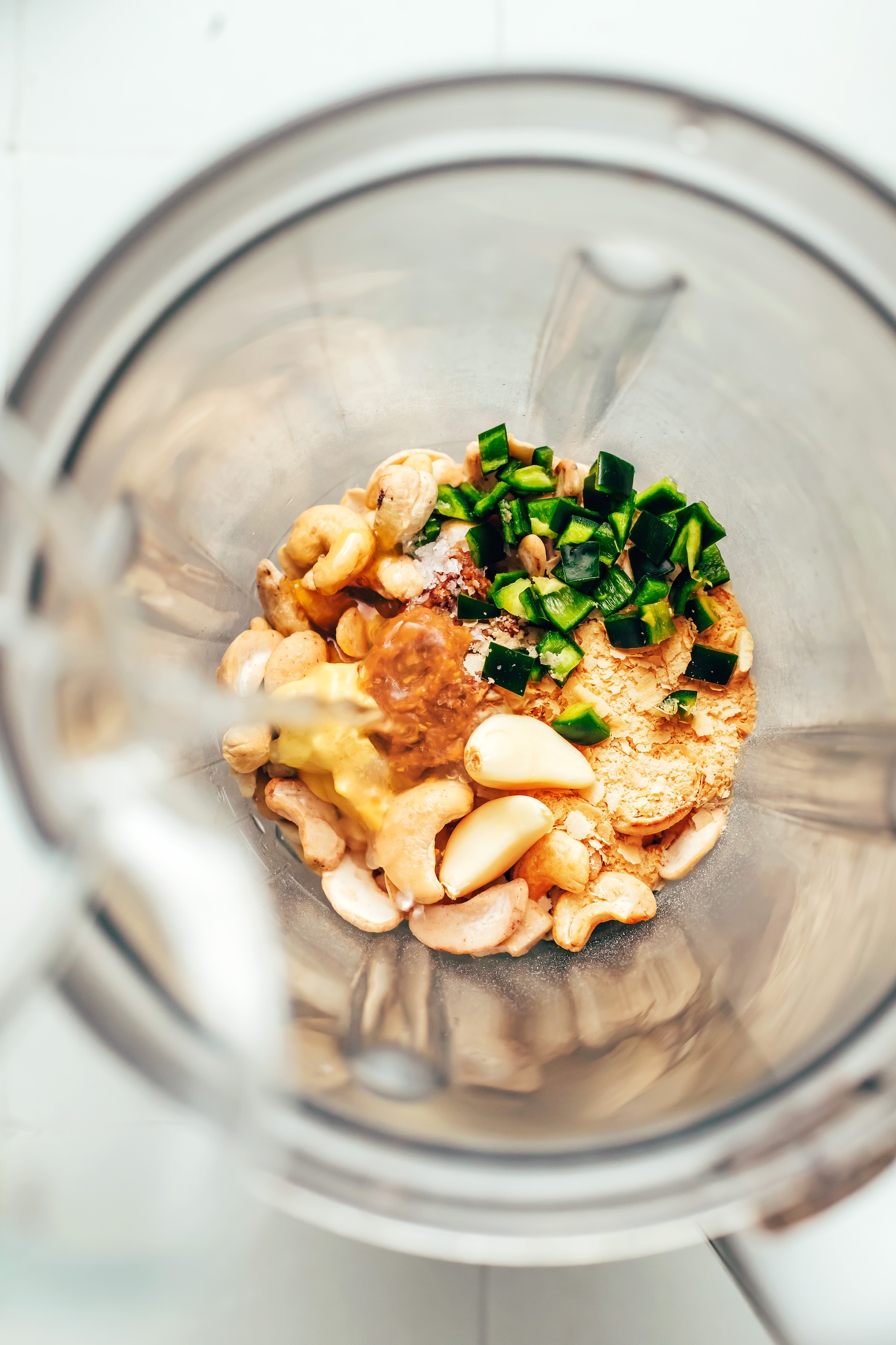 Pouring water into a blender with cashews, lime juice, nutritional yeast, miso paste, Dijon mustard, maple syrup, salt, garlic, and chopped jalapeño